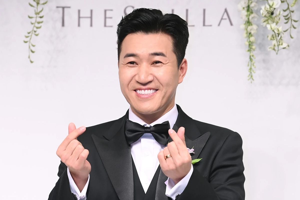 김종민이 검은색 정장을 입고 손가락 하트를 하고 있다.