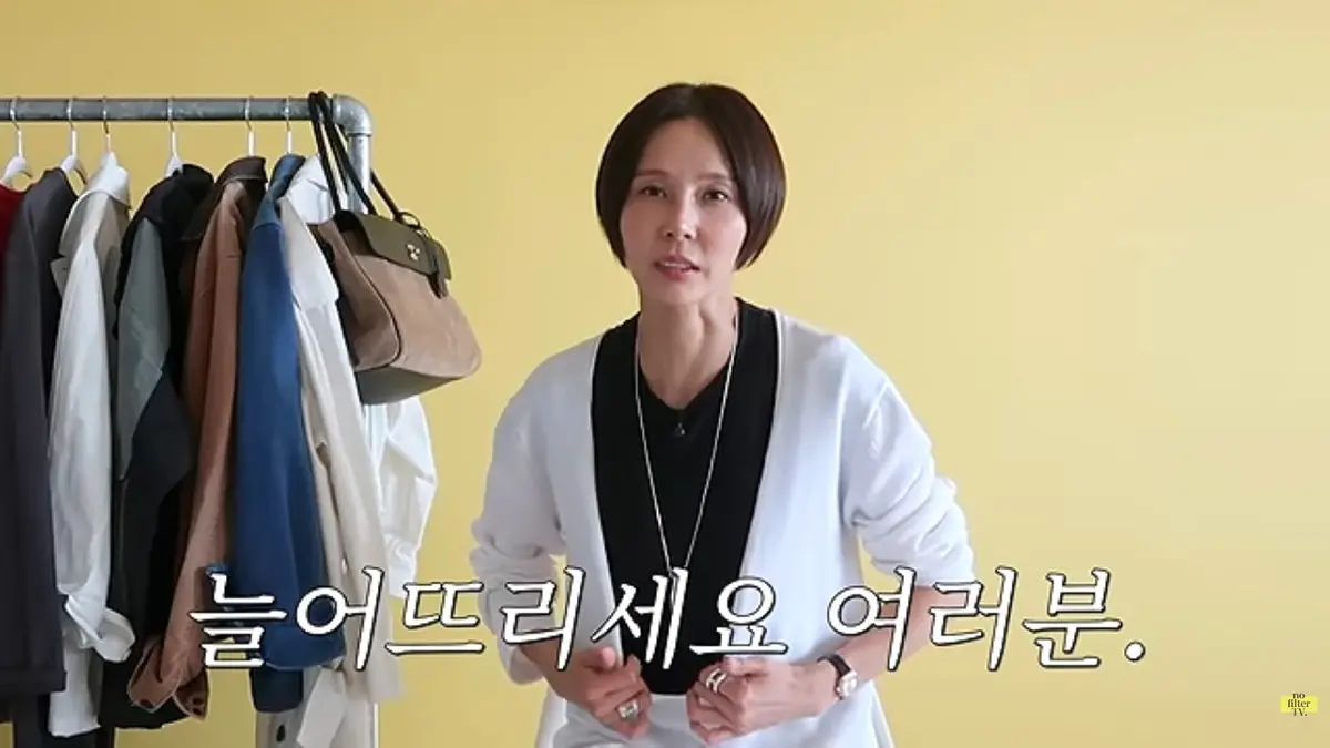 옷걸이에 걸린 다양한 옷들 앞에서 흰색 가디건을 입고 있는 여성.