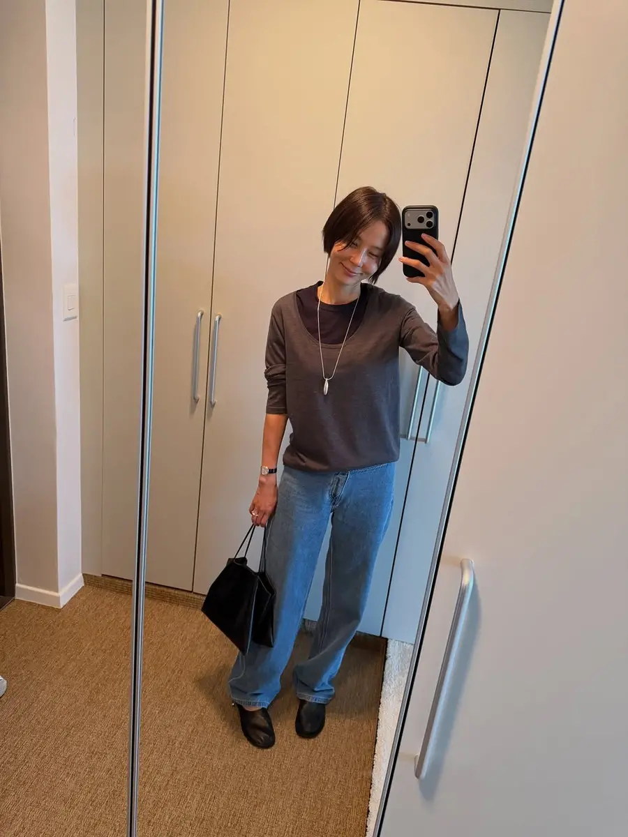 거울 앞에서 회색 상의와 청바지를 입고 셀카를 찍는 여성.