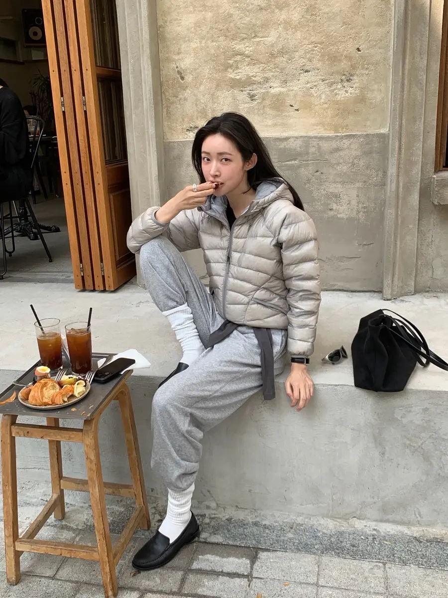 회색 경량 패딩과 트레이닝복을 입은 여성이 야외에 앉아 있다.